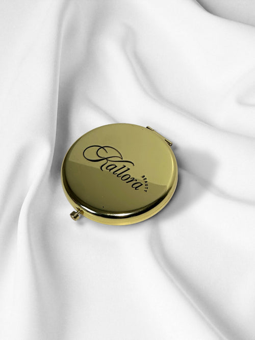 Gold compact mirror with 'Kallora' branding on a white fabric background